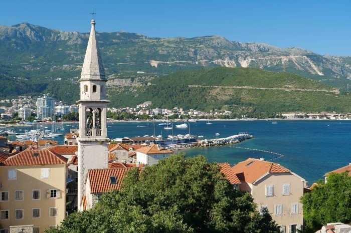 La aguja de la iglesia se eleva sobre edificios beige en Budka Montenegro con puerto, Barcos y altos acantilados verdes en segundo plano.