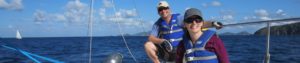 Close-up of a guy sitting behind a woman steering a Colgate 26, both wearing blue life vests, learning to sail in British Virgin Islands