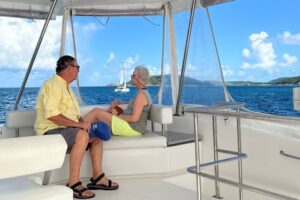 A couple relaxing on a bench on the top level of a 52' power catamaran with a catamaran under sail seen in the distance between them