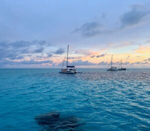 Dark reef in clear waters with boats anchored behind